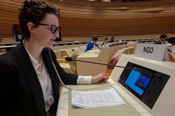 [ai] A woman wearing glasses sits at a conference table, speaking into a microphone. A sign labeled 'NGO' is visible beside her. Participants are seated in the background, engaged in the conference.
